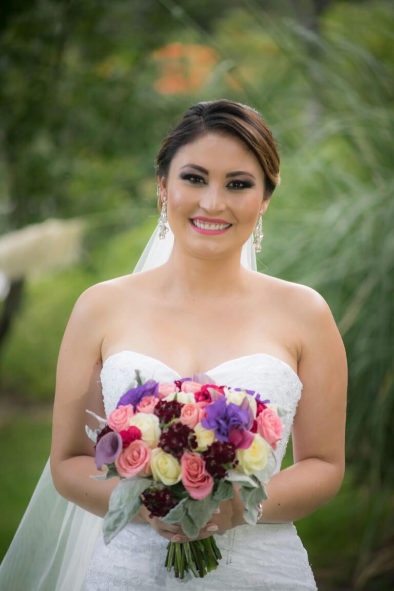 A cheerful bride in a white dress holding a vibrant bouquet outdoors, exuding happiness and elegance.
